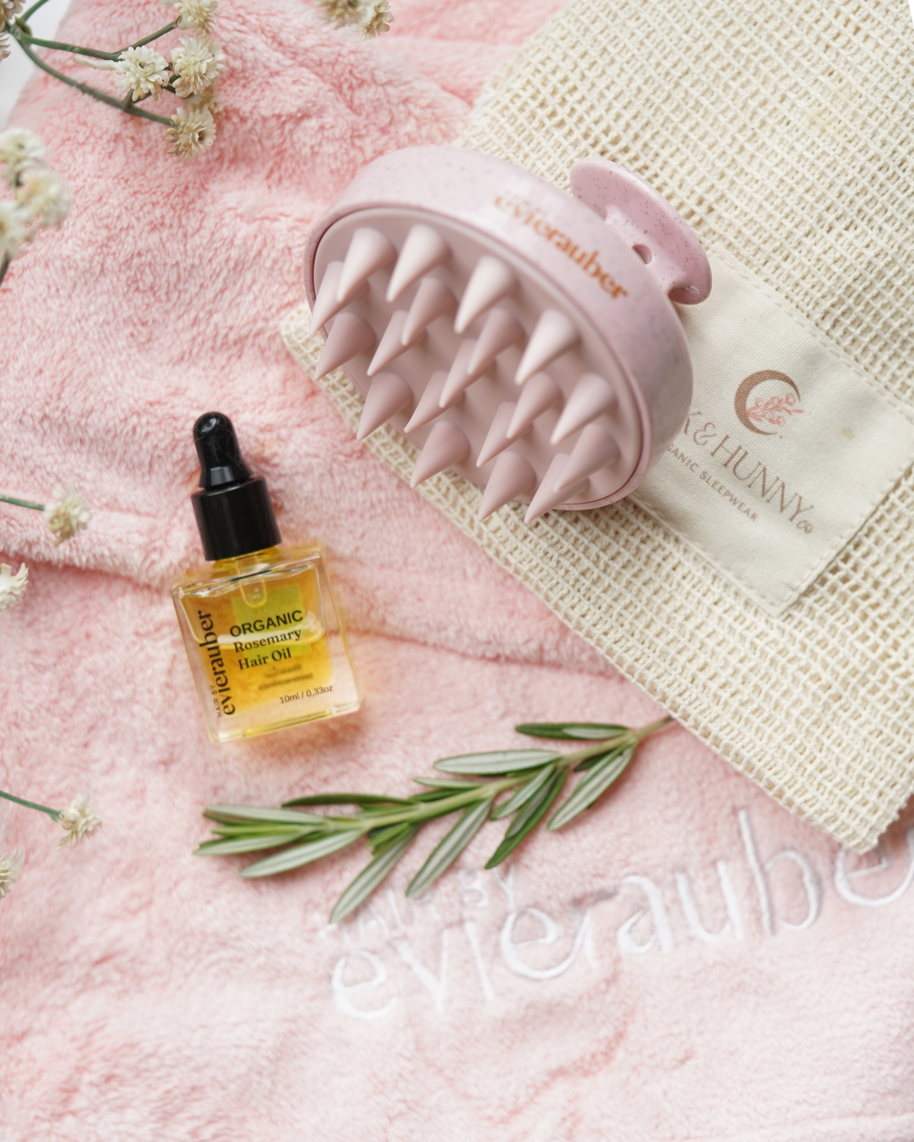 A pink scalp Brush, displayed on a cotton laundry bag l alongside a bottle of evie rauber Organic Nourishing Hair Oil and rosemary sprigs on a pink towel.