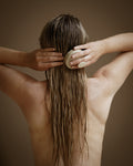 A woman with long, wet brown hair seen from behind, holding a beige scalp brush against the back of her head with both hands, set against a solid warm brown background.