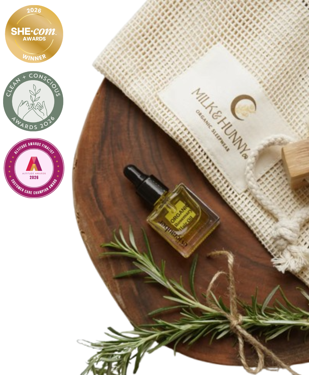 A styled product shot featuring a small amber glass dropper bottle of essential oil with a black cap, placed on a round wooden board next to a cream mesh drawstring pouch labeled 'MILK & HUNNY' and a sprig of rosemary. Three award badges are shown along the left side on a white background.