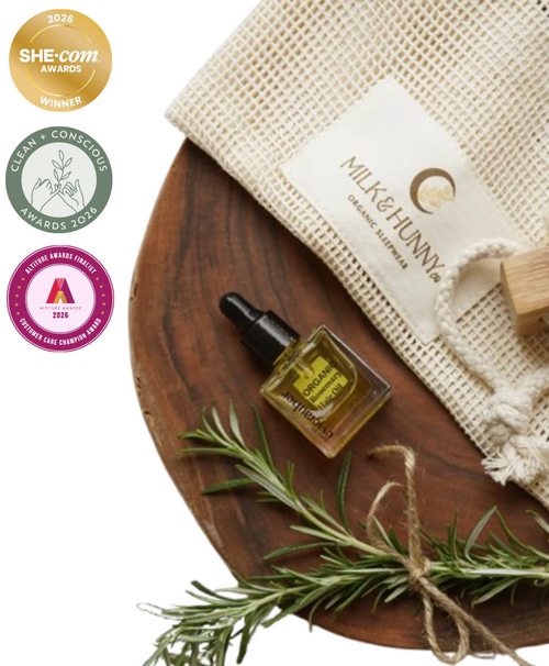 A styled product shot featuring a small amber glass dropper bottle of essential oil with a black cap, placed on a round wooden board next to a cream mesh drawstring pouch labeled 'MILK & HUNNY' and a sprig of rosemary. Three award badges are shown along the left side on a white background.