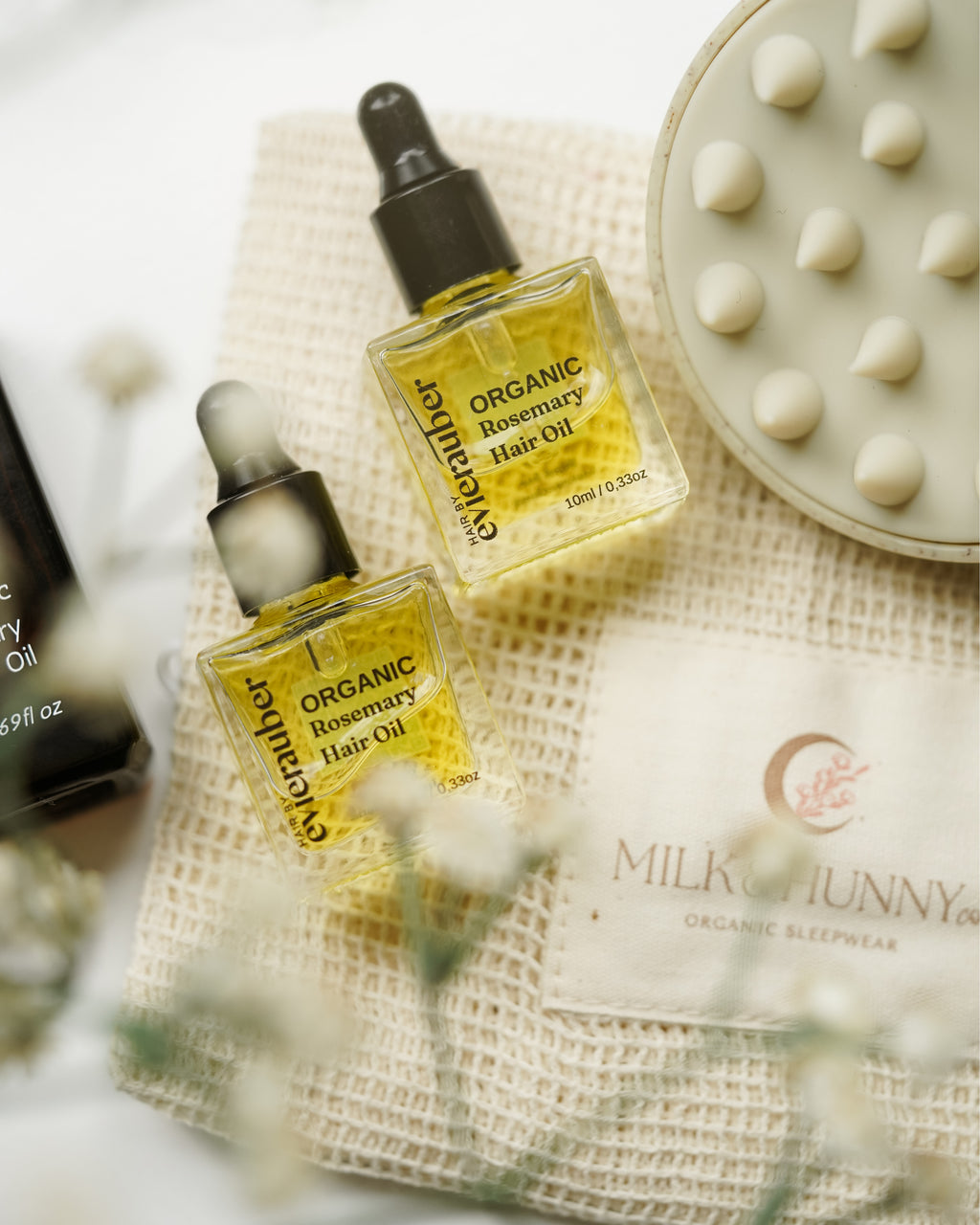 Two bottles of Organic Rosemary Hair Oil and a laundry bag are displayed on a textured cream fabric alongside a spa-style silicone scalp massager and a blurred bottle in the background.